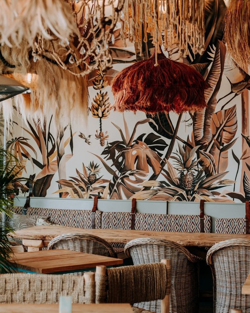 El interior de Petit Nola: un mural de selva pintado a mano, lámparas de rafia y macramé, bancos con cojines de kilim y sillas de mimbre.