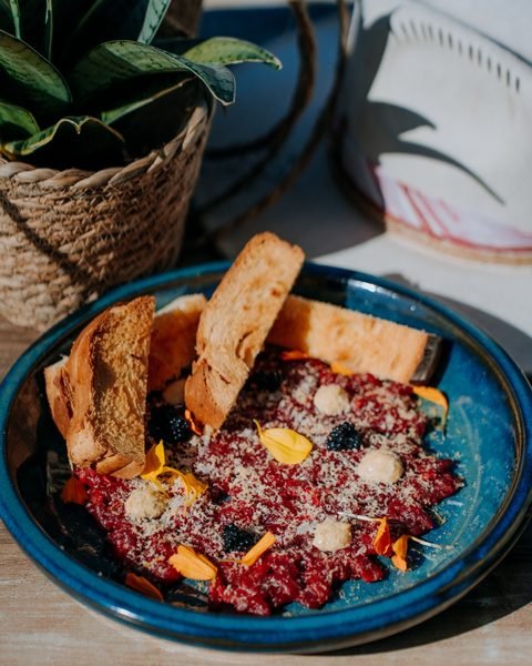 Steak tartar trufado servido en un plato de cerámica azul con brioche tostado y flores comestibles.