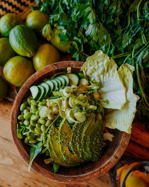 El bol Diosa Verde: aguacate, edamame, pepino y brotes en un cuenco de madera, rodeado de limas y hierbas frescas.